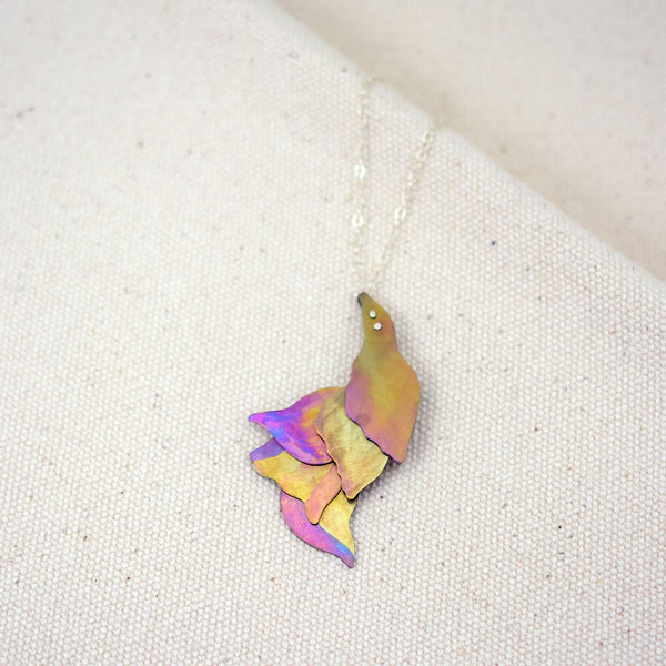 detail of She Wades with Grace Through Changing Tides Necklace pictured on a white woven fabric, a pink and gold hand-painted titanium necklace of layered organic shapes similar to flower petals or feathers