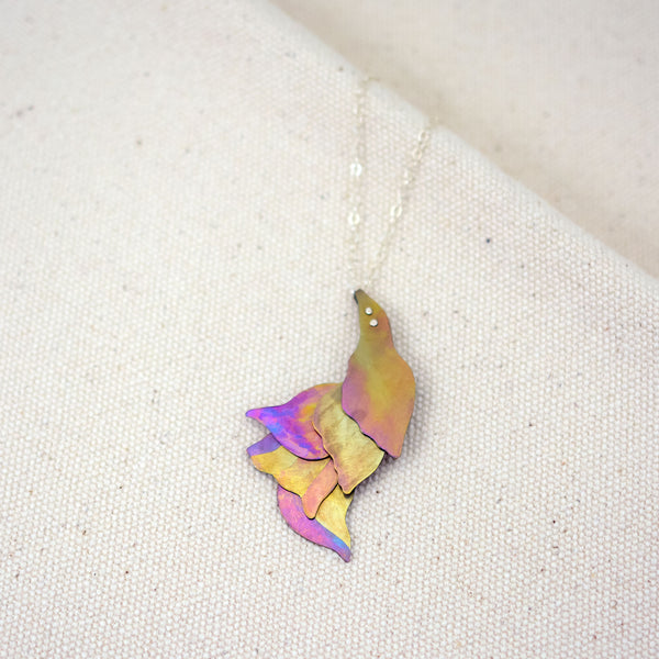 detail of She Wades with Grace Through Changing Tides Necklace pictured on a white woven fabric, a pink and gold hand-painted titanium necklace of layered organic shapes similar to flower petals or feathers
