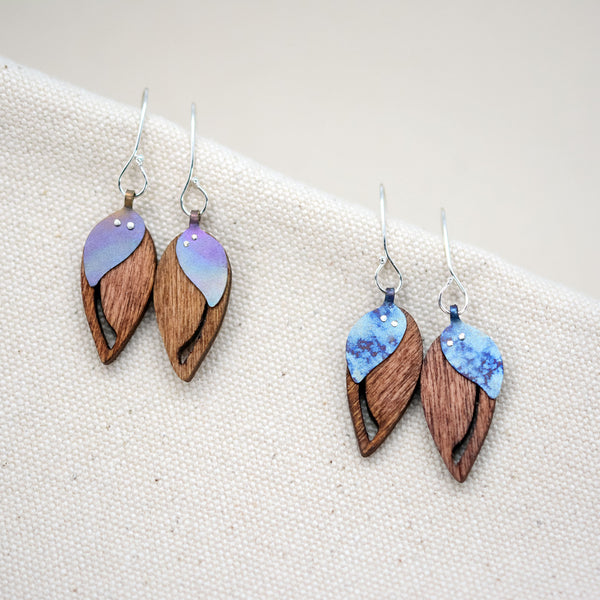 the two colorful hand painted patterns of The Dew at Dawn earrings, upside-down teardrop-shaped wooden earrings with a slit down the side and a blue teardrop-shaped piece of titanium riveted on top