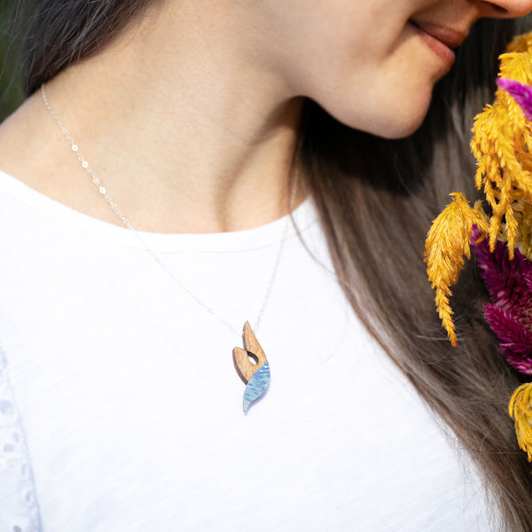 woman's neckline modeling The Wind in Her Sails necklace, a wooden and titanium organic swoop shaped curl hand painted in blue and yellow